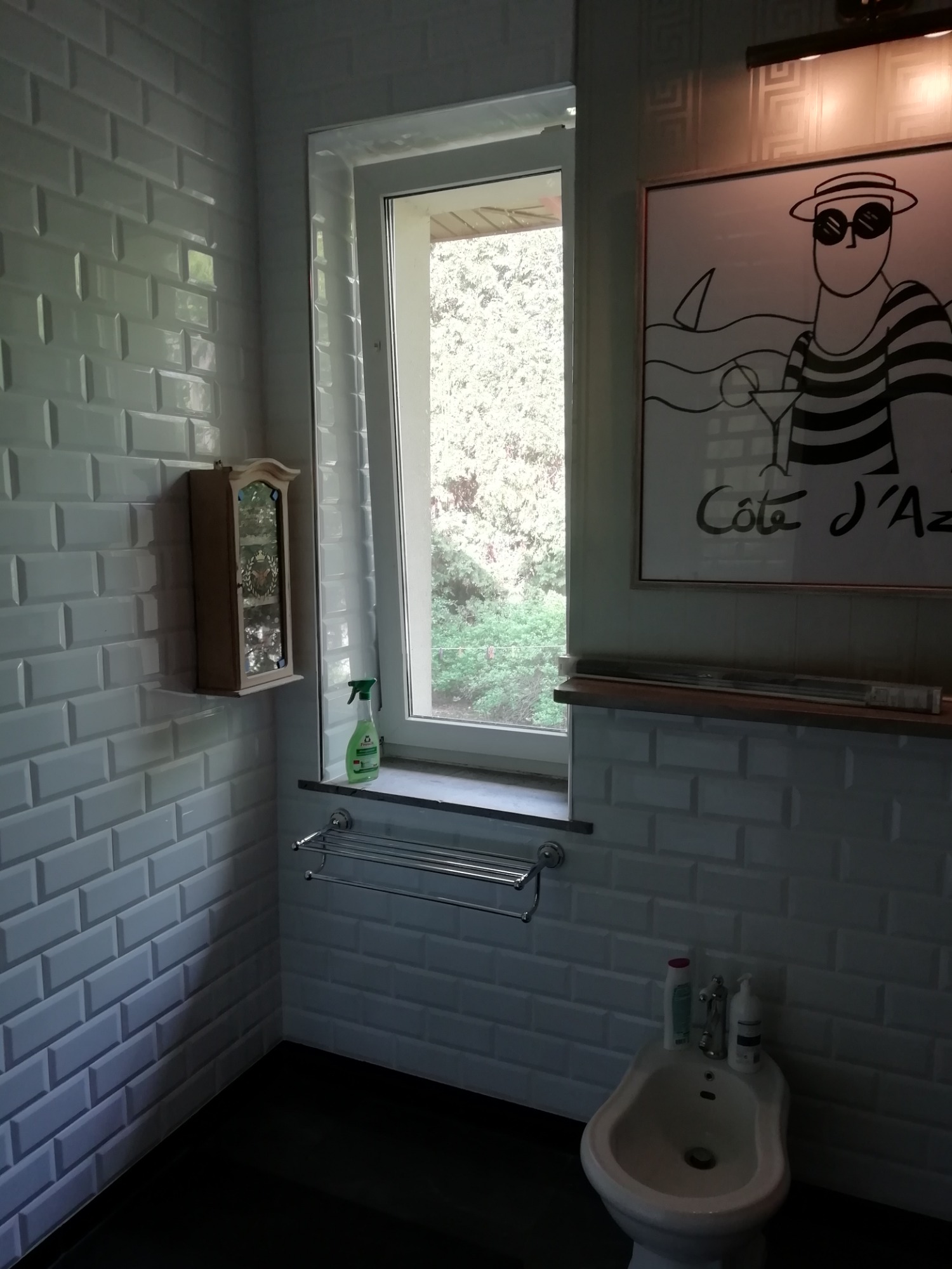 Wykończona łazienka z białą, cegiełkową glazurą, bidetem, szafką wiszącą i oknem z widokiem na zieleń, ozdobiona obrazkiem w stylu retro z napisem 'Cote d'Azur'.