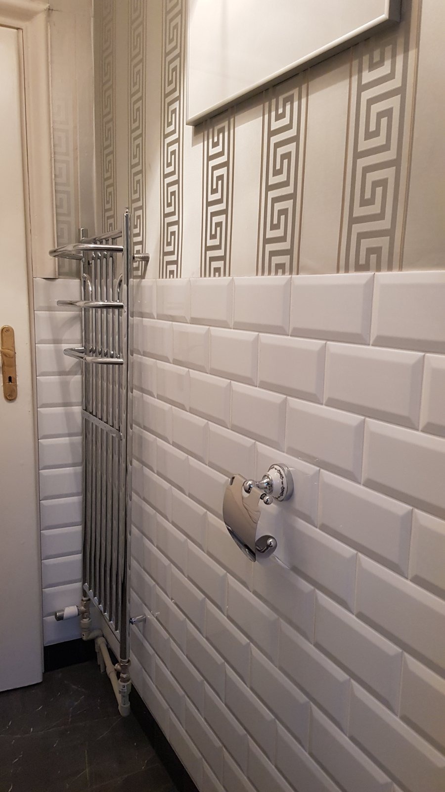 Białe, fazowane płytki ścienne typu metro w łazience, z chromowanym grzejnikiem drabinkowym i uchwytem na papier toaletowy z porcelanowymi akcentami, na tle tapety w geometryczne wzory.