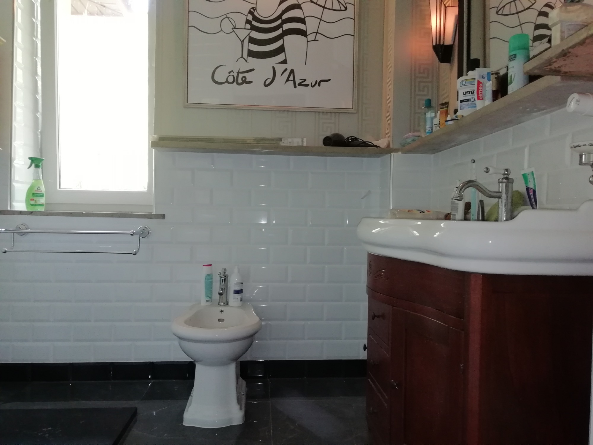 Biała łazienka z bidetem, umywalką z szafką z ciemnego drewna i białymi płytkami metra, ozdobiona grafiką z motywem Côte d'Azur.