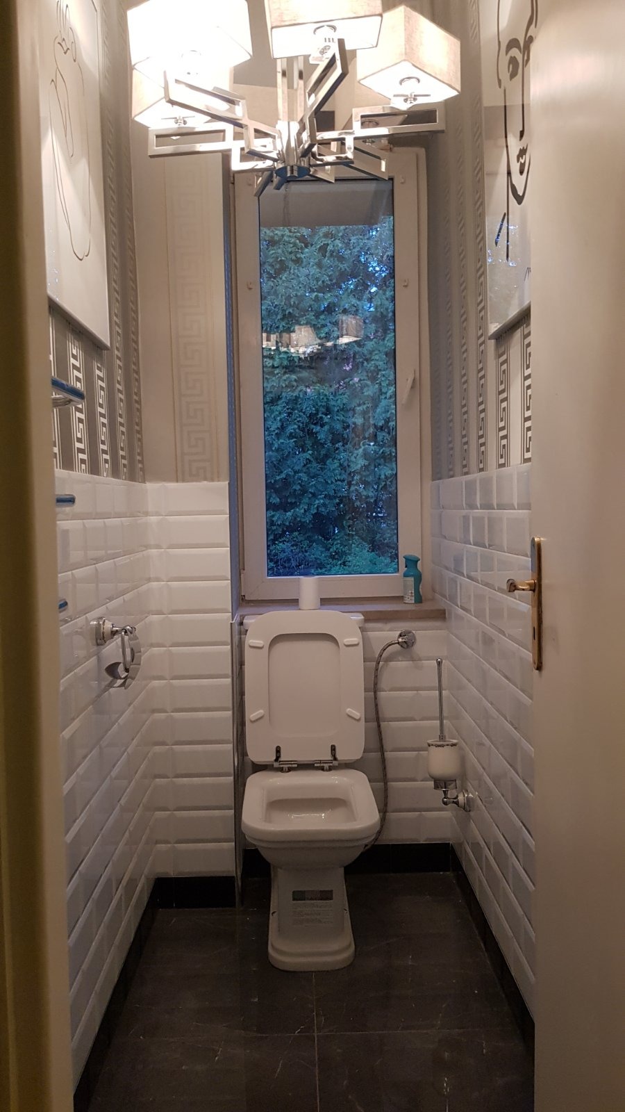 Wąska toaleta z białymi płytkami typu metro, ciemną podłogą i oknem z widokiem na zieleń, oświetlona nowoczesnym żyrandolem i ozdobiona grafiką na ścianie.