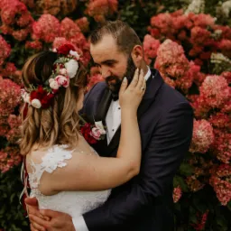 Para młoda w objęciach na tle kwitnących hortensji: panna młoda w wianku z czerwonych i białych kwiatów, pan młody w granatowym smokingu.