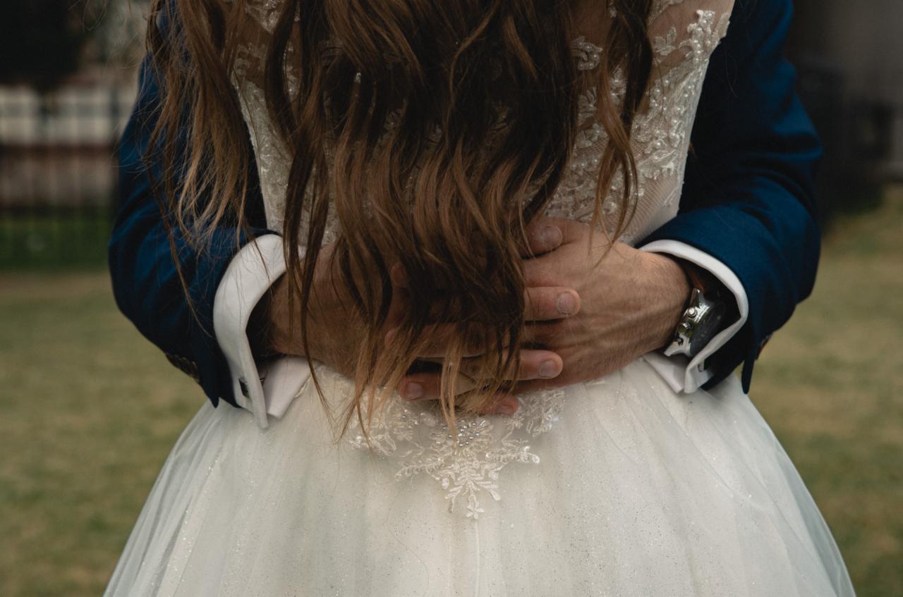 Mężczyzna w granatowym garniturze obejmuje kobietę w białej sukni ślubnej, widoczne detale sukni i zegarek na ręce mężczyzny.