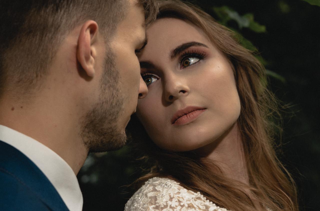 Zbliżenie na parę: kobieta w koronkowej sukni z makijażem i mężczyzna w niebieskiej marynarce, delikatny kontakt twarzami na tle zieleni.