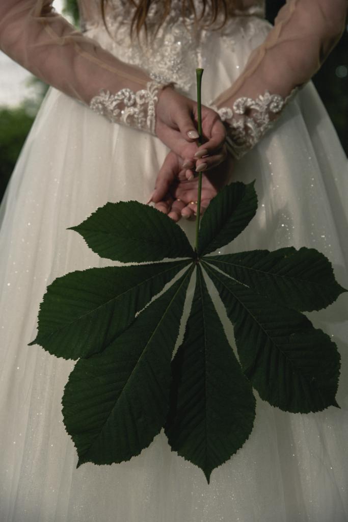 Panna młoda w sukni ślubnej z koronkowymi rękawami trzyma przed sobą duży, zielony liść kasztanowca.