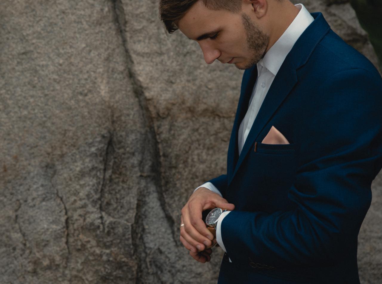 Mężczyzna w granatowym garniturze, z poszetką i zegarkiem na tle skalnej ściany, spogląda na zegarek.
