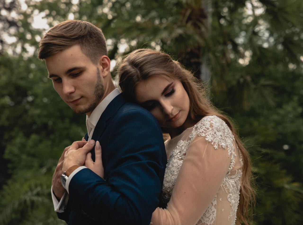 Młoda para w objęciach na tle zieleni: panna młoda w koronkowej sukni opiera głowę na ramieniu pana młodego w granatowym garniturze, delikatne światło podkreśla makijaż panny młodej.