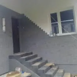 Elewacja domu z szarej cegły klinkierowej, schody wejściowe wyłożone tym samym materiałem, widoczne okno z białymi ramami i lampa zewnętrzna przy drzwiach.