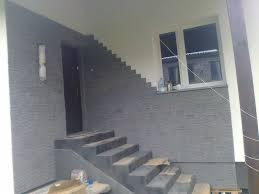 Elewacja domu z szarej cegły klinkierowej, schody wejściowe wyłożone tym samym materiałem, widoczne okno z białymi ramami i lampa zewnętrzna przy drzwiach.