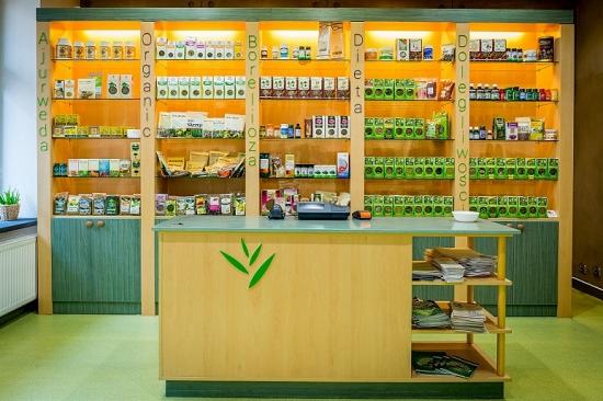 Sklep z ziołami i suplementami diety: regały z produktami oznaczonymi kategoriami Ajurweda, Organic, Borelioza, Dieta i Dolegliwości, kasa samoobsługowa z zielonym logo liści na ladzie.