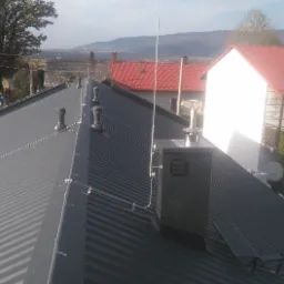 Szary blaszany dach z zamontowanym systemem odgromowym, kominami wentylacyjnymi i metalową obudową kominową. W tle zabudowa miejska i zalesione wzgórza.