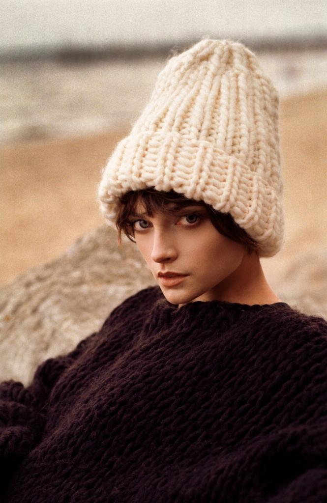 Portret kobiety w wełnianej czapce z grubym splotem i ciemnym swetrze na tle plaży z widocznymi kamieniami i piaskiem.