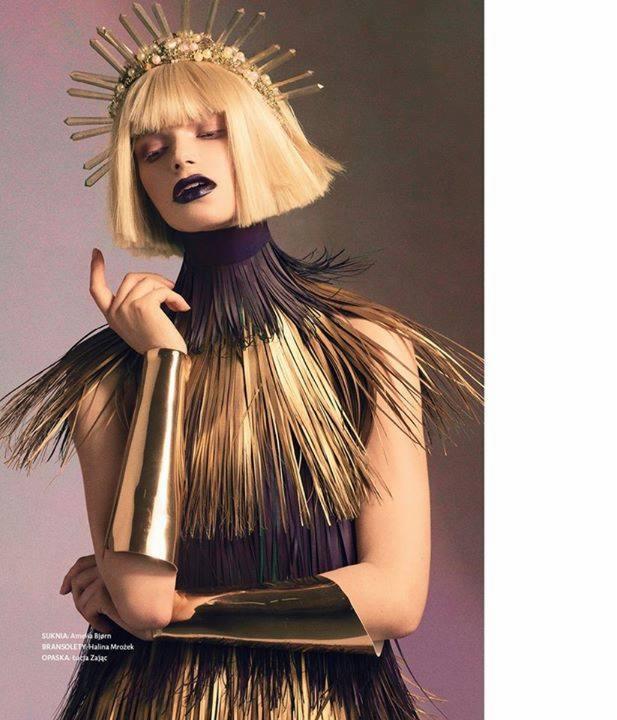 Kobieta z blond bobem, w koronie z promieniami i sukni z metalicznymi frędzlami, pozująca z uniesioną dłonią i ciemną szminką.