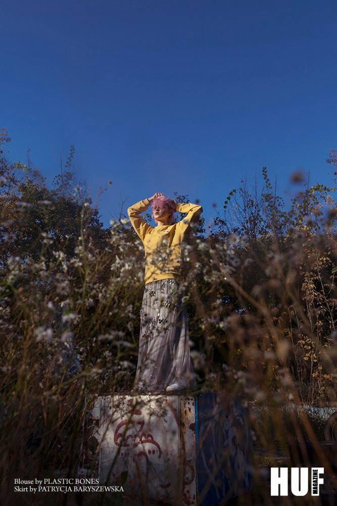 Kobieta z różowymi włosami w żółtej bluzie i długiej, zdobionej spódnicy pozuje na betonowym cokole wśród wysokich traw i kwiatów na tle błękitnego nieba, ujęcie z dołu.