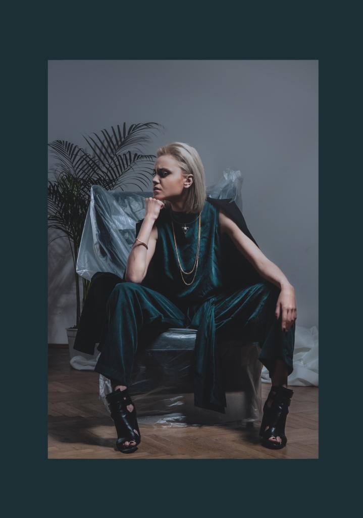 Blondwłosa modelka w szmaragdowym welurowym kombinezonie i czarnych botkach na obcasie siedzi na krześle obłożonym folią, zamyślona, z dłonią pod brodą, w tle roślina doniczkowa i biała tkanina.