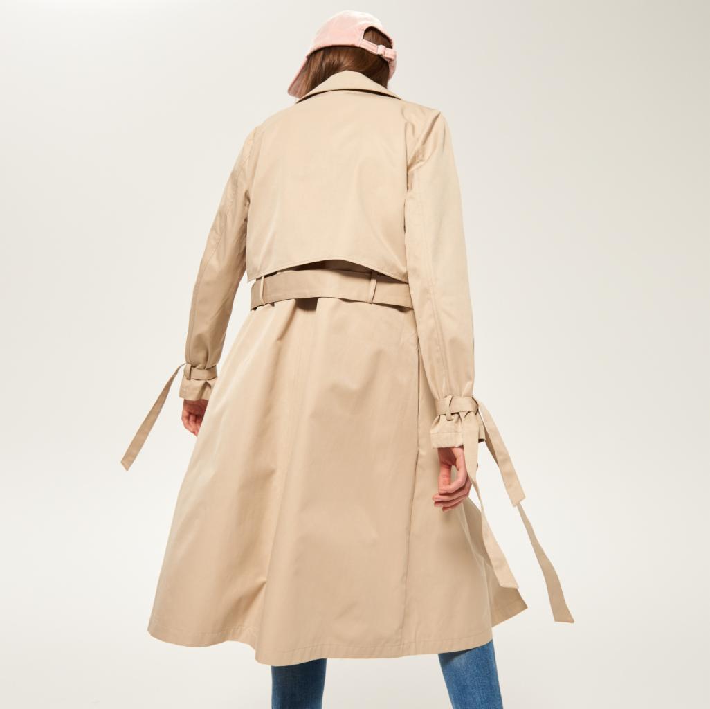 Beżowy trencz damski z paskiem w talii i wiązaniami na rękawach, widziany od tyłu, z różową czapką bejsbolówką na głowie modelki.