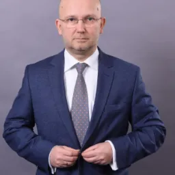 Mężczyzna w niebieskim garniturze zapina guzik marynarki, patrząc wprost, na szarym tle. Widoczne okulary i krawat w kropki.