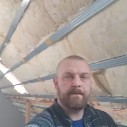 Mężczyzna z brodą na tle ocieplonego poddasza z widoczną wełną mineralną i metalową konstrukcją nośną