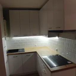 Nowoczesna, biała kuchnia narożna z drewnianym blatem, oświetleniem podszafkowym i płytkami typu metro na ścianie.