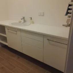 Biała szafka łazienkowa na wymiar z umywalką nablatową, chromowaną armaturą i drewnopodobną podłogą z płytek.