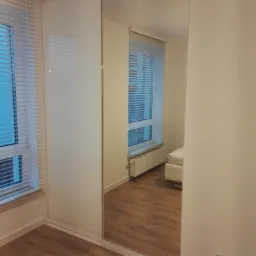 Biała szafa wnękowa z przesuwnymi drzwiami, jedno skrzydło z matowego szkła, drugie lustrzane, odbijające wnętrze pokoju z oknem i grzejnikiem.