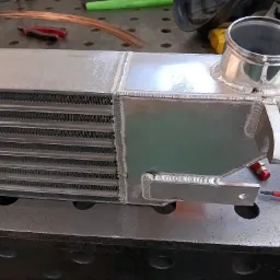 Posrebrzany intercooler po spawaniu, z widocznymi spawami, umieszczony na metalowym stole spawalniczym. W tle narzędzia i drut spawalniczy.