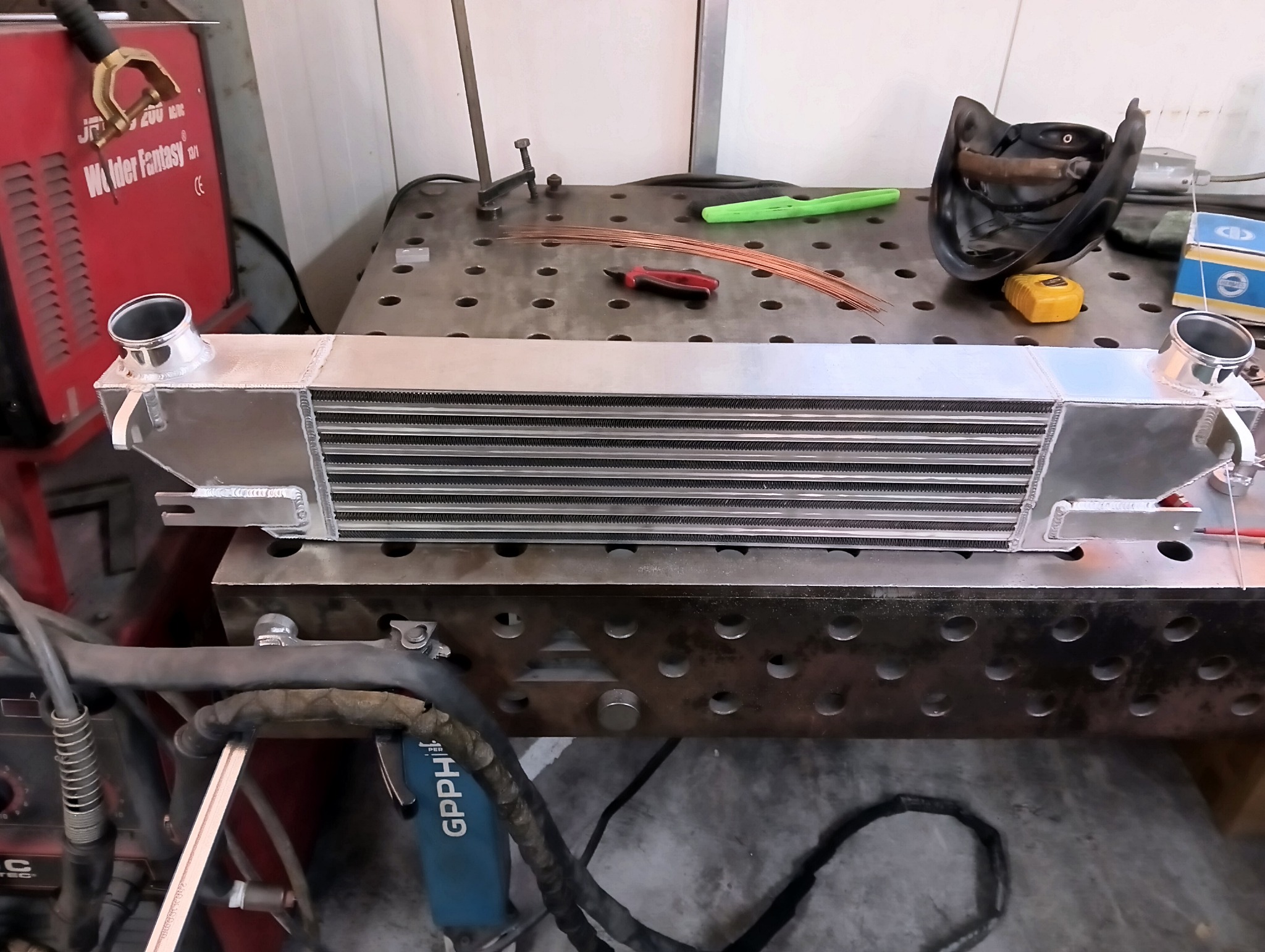 Aluminiowy intercooler na perforowanym stole spawalniczym, widoczny sprzęt spawalniczy i maska. Detaliczna obróbka metalu, precyzyjne spawy.