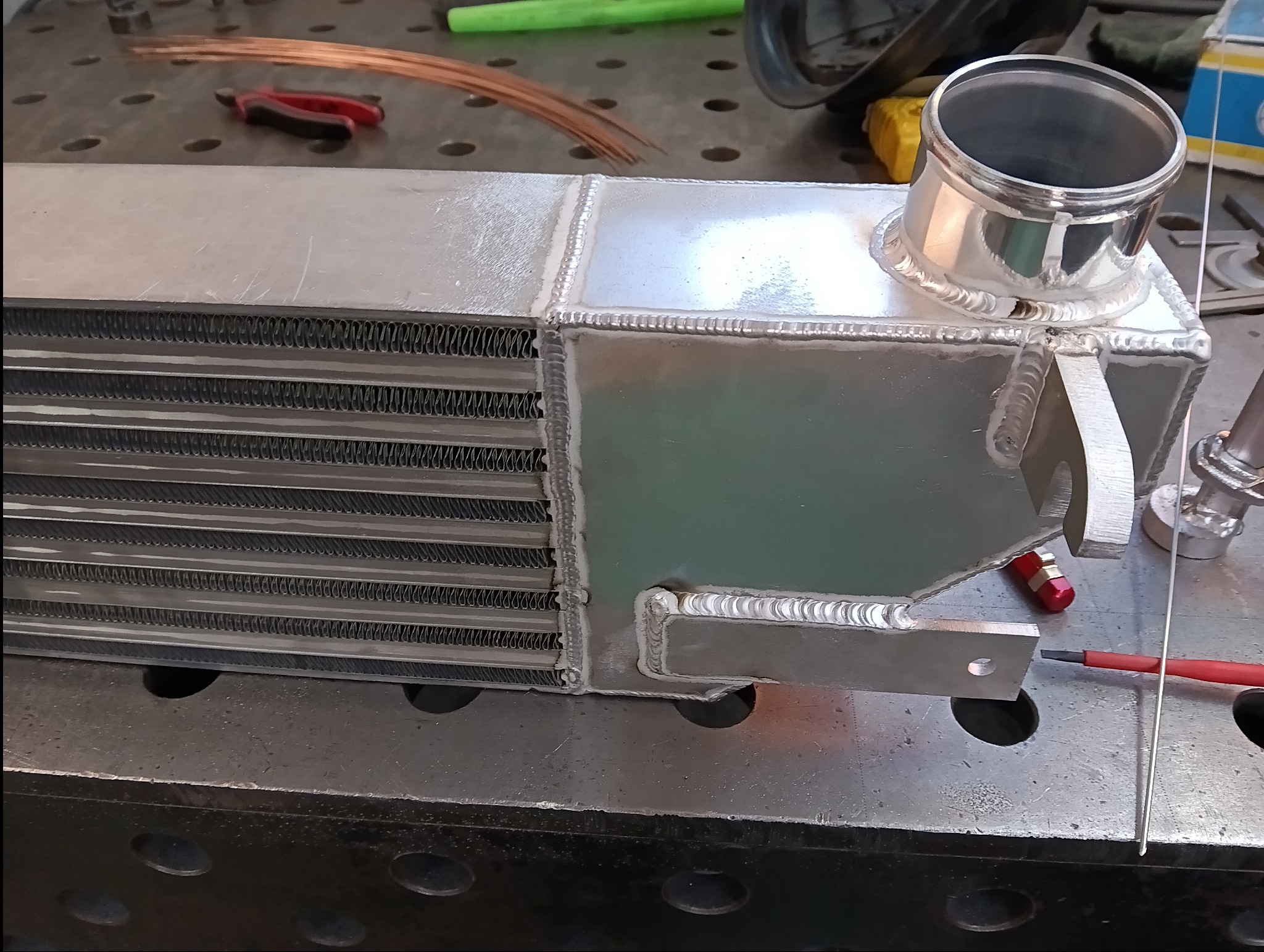 Posrebrzany intercooler po spawaniu, z widocznymi spawami, umieszczony na metalowym stole spawalniczym. W tle narzędzia i drut spawalniczy.
