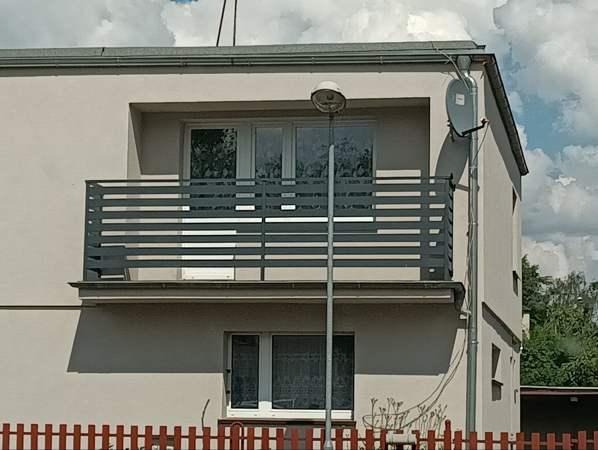 Szary balkon z poziomymi szczeblami przy jasnym budynku. Na elewacji widoczna antena satelitarna. W tle błękitne niebo z chmurami.