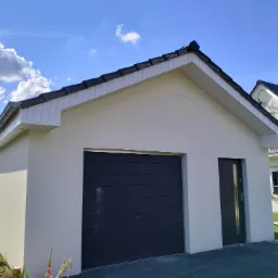 Nowo otynkowany garaż z ciemną bramą segmentową i drzwiami bocznymi, biała podbitka dachowa, widoczne fragmenty domu z ciemną dachówką w tle, błękitne niebo z chmurami.