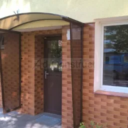 Zadaszenie nad wejściem do budynku z brązową ramą i przezroczystym daszkiem, obok białe okno z widocznym odbiciem samochodu.