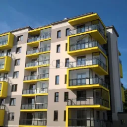 Nowoczesny budynek wielorodzinny w Sulejówku z przeszklonymi balkonami o żółtych ramach, widok z dołu w słoneczny dzień.