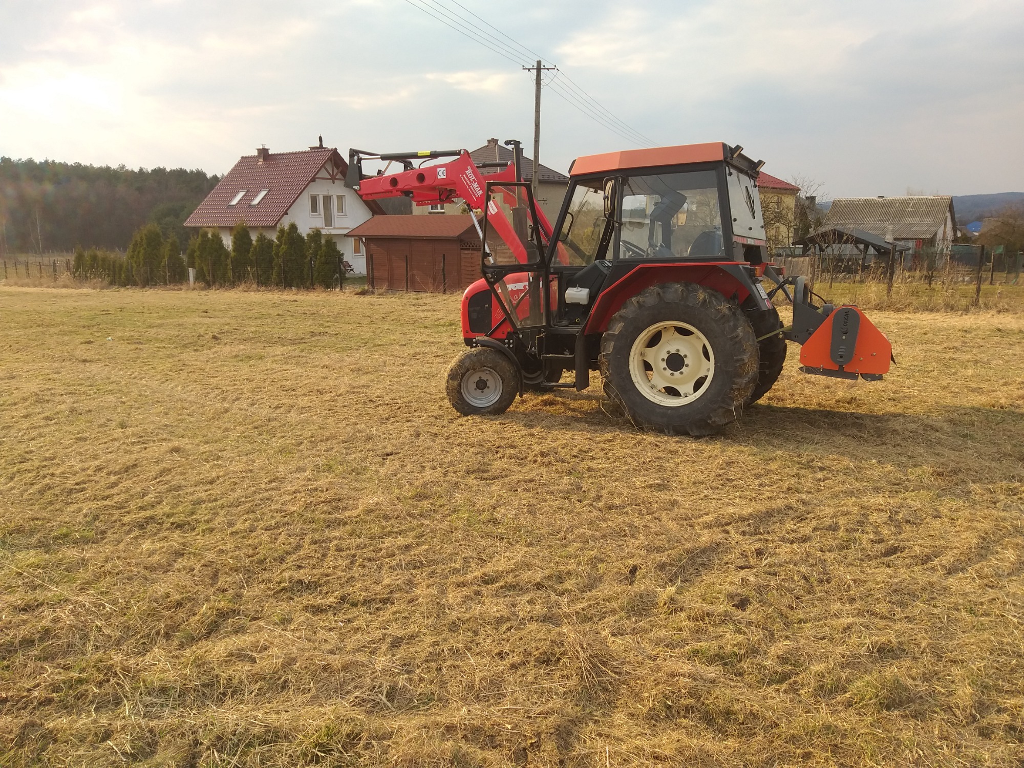 Czerwony traktor z zamontowanym urządzeniem tnącym na polu z niską, wysuszoną trawą, w tle domy i drzewa.