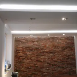 Wnętrze pokoju z ceglaną ścianą, podwieszanym sufitem z oświetleniem LED, widoczne narzędzia i materiały budowlane.