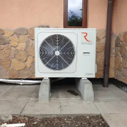 Biała jednostka zewnętrzna klimatyzacji marki Rotenso Aquami Series, zamontowana na betonowych wspornikach przed budynkiem z kamienną elewacją i brązowym oknem.