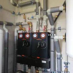 Rozbudowana instalacja grzewcza z licznymi rurami, zaworami, dwoma czarnymi modułami marki Termojet z manometrami, izolacją termiczną na rurach i białym zasobnikiem ciepłej wody.
