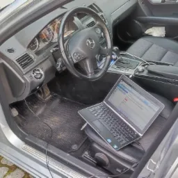 Wnętrze srebrnego samochodu Mercedes-Benz z otwartymi drzwiami; na siedzeniu pasażera leży otwarty laptop z widocznym interfejsem diagnostycznym.