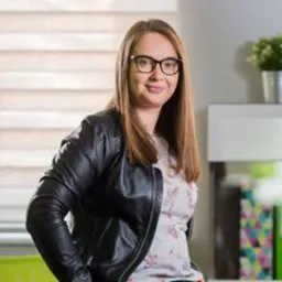 Uśmiechnięta kobieta w okularach i czarnej kurtce skórzanej, z długimi brązowymi włosami, pozuje na tle jasnego biura z rośliną doniczkową i kolorowymi książkami.