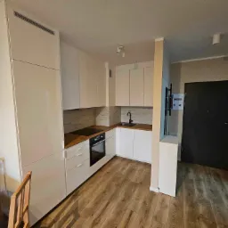 Nowoczesna, jasna kuchnia z białymi szafkami, drewnianym blatem i podłogą, widziana z perspektywy jadalni z drewnianym stołem i krzesłami.