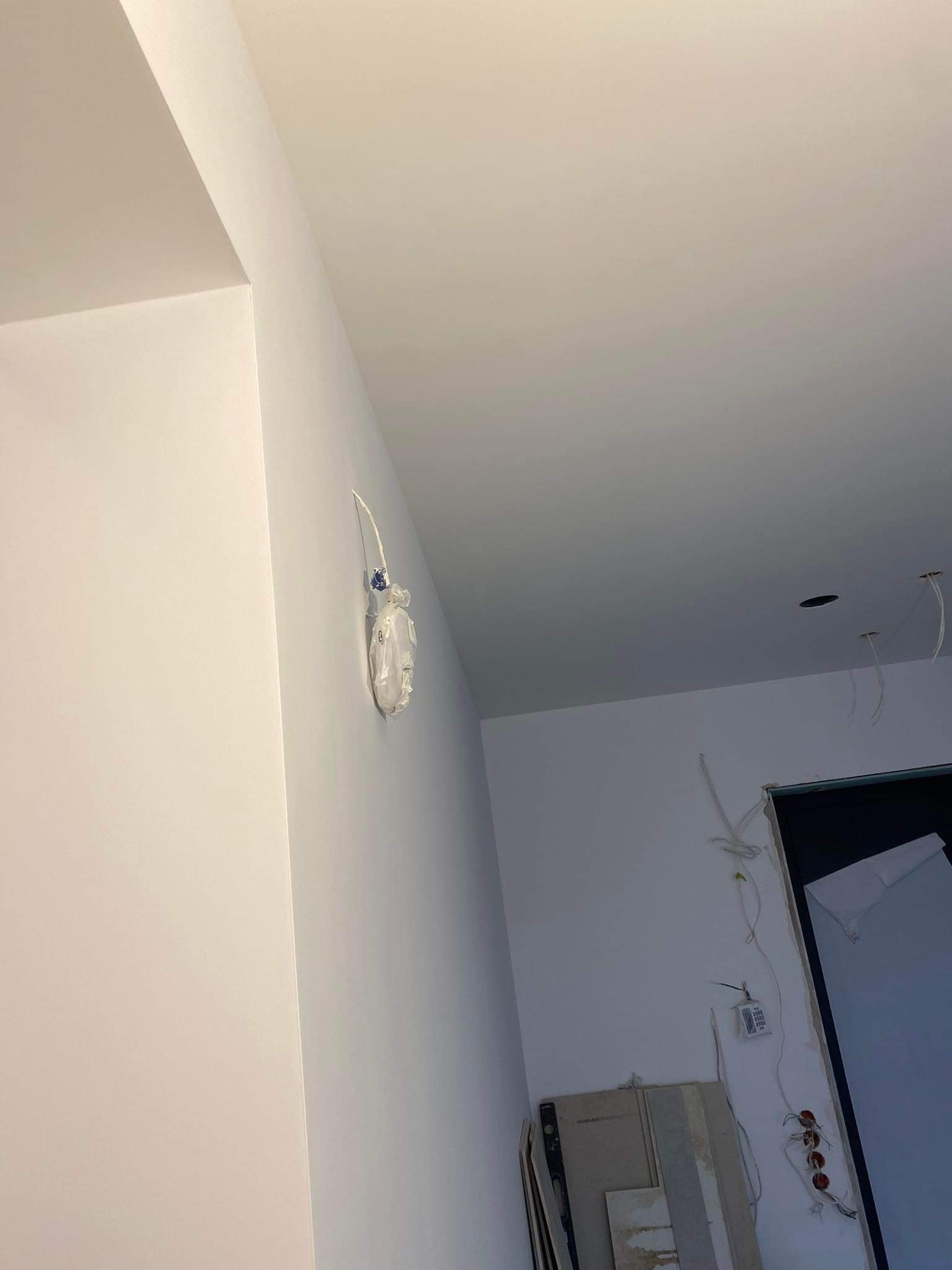 Świeżo pomalowane, gładkie ściany w jasnym odcieniu z zabezpieczonym gniazdkiem elektrycznym oraz wystającymi przewodami, widoczne przygotowania do dalszych prac wykończeniowych.