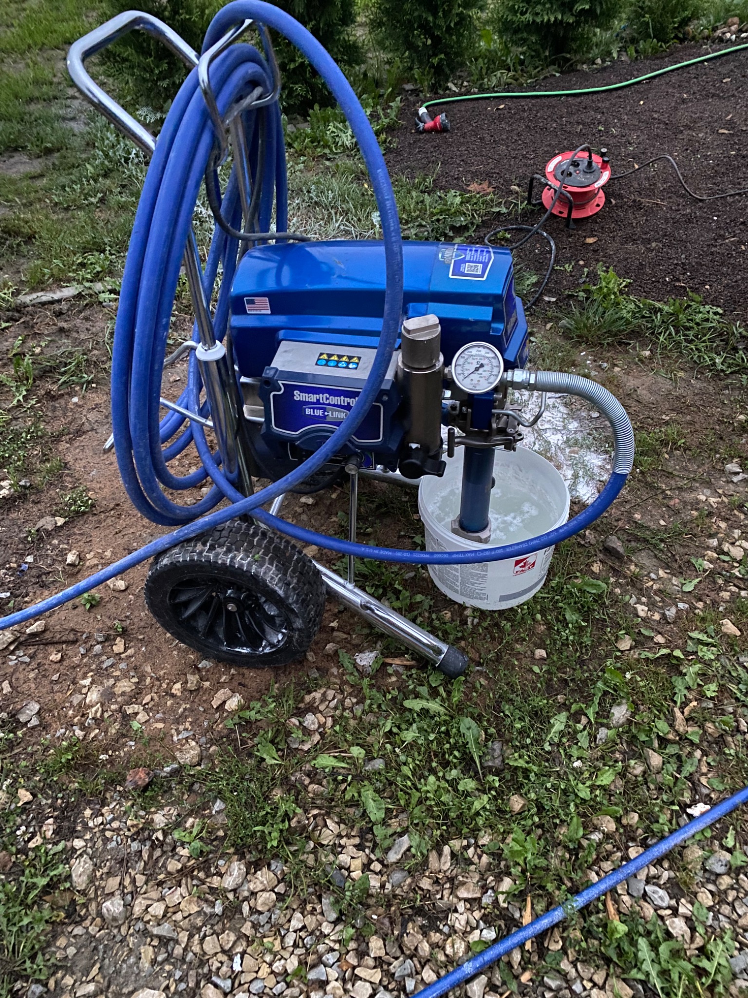Profesjonalny, mobilny agregat malarski w kolorze niebieskim z manometrem i wężem zanurzonym w wiadrze z wodą, na tle ogrodu z rozłożonym przedłużaczem.