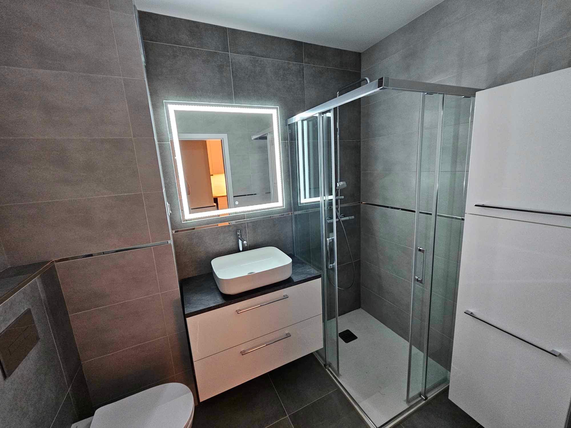 Nowoczesna łazienka z szarymi płytkami, podświetlanym lustrem, białą umywalką nablatową na szafce z szufladami i kabiną prysznicową z przesuwnymi drzwiami.