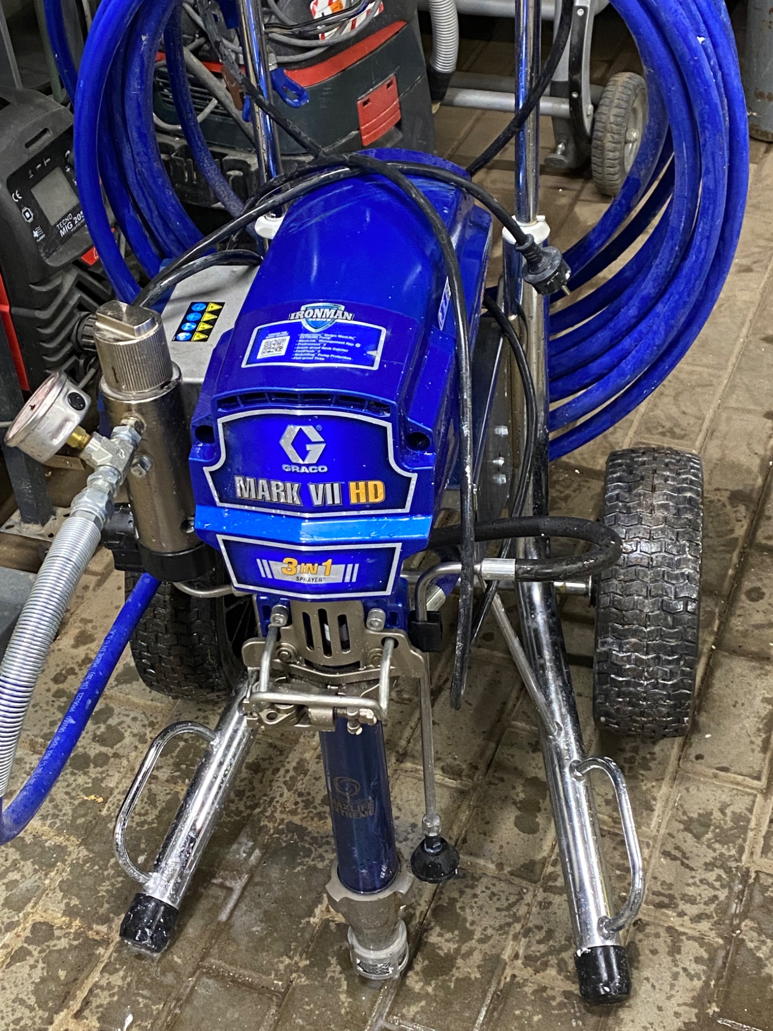Profesjonalny agregat malarski Graco Mark VII HD na kółkach z podłączonym niebieskim wężem, widoczny manometr i elementy sterujące, stojący na brudnej, kafelkowanej podłodze w warsztacie.