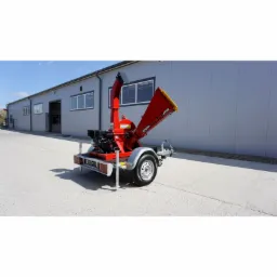 Czerwony rębak spalinowy tarczowy RTS-630 na przyczepie, widoczny na betonowym placu przed szarym budynkiem magazynowym w słoneczny dzień.