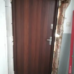 Brązowe drzwi wewnętrzne z ościeżnicą, zamontowane w surowym otworze z widocznymi fragmentami cegieł i resztkami zaprawy, poziomica oparta o ścianę w tle.