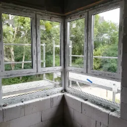 Narożne okno PCV z widokiem na rusztowania i zieleń, w trakcie montażu w surowym murze z betonu komórkowego, widoczne ślady pianki montażowej.