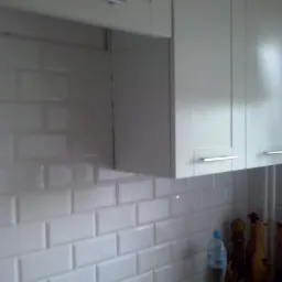 Jasne, wiszące szafki kuchenne z poziomymi uchwytami nad białą, cegiełkową glazurą, widoczny fragment blatu z butelką wody i akcesoriami.