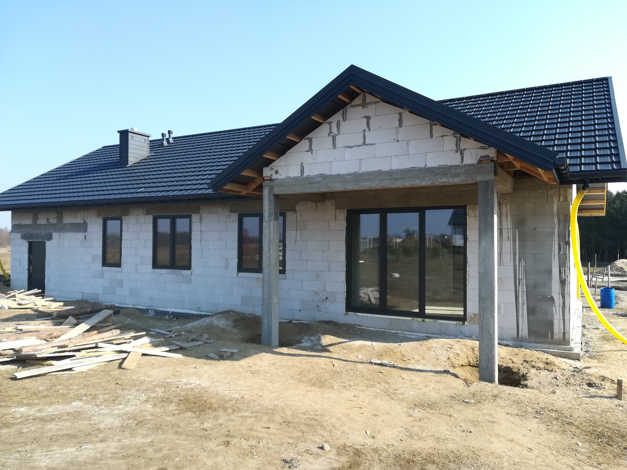 Dom w stanie surowym otwartym z zamontowanymi oknami PCV w czarnych ramach, widoczne drewniane deski na ziemi i żółty wąż doprowadzający media.