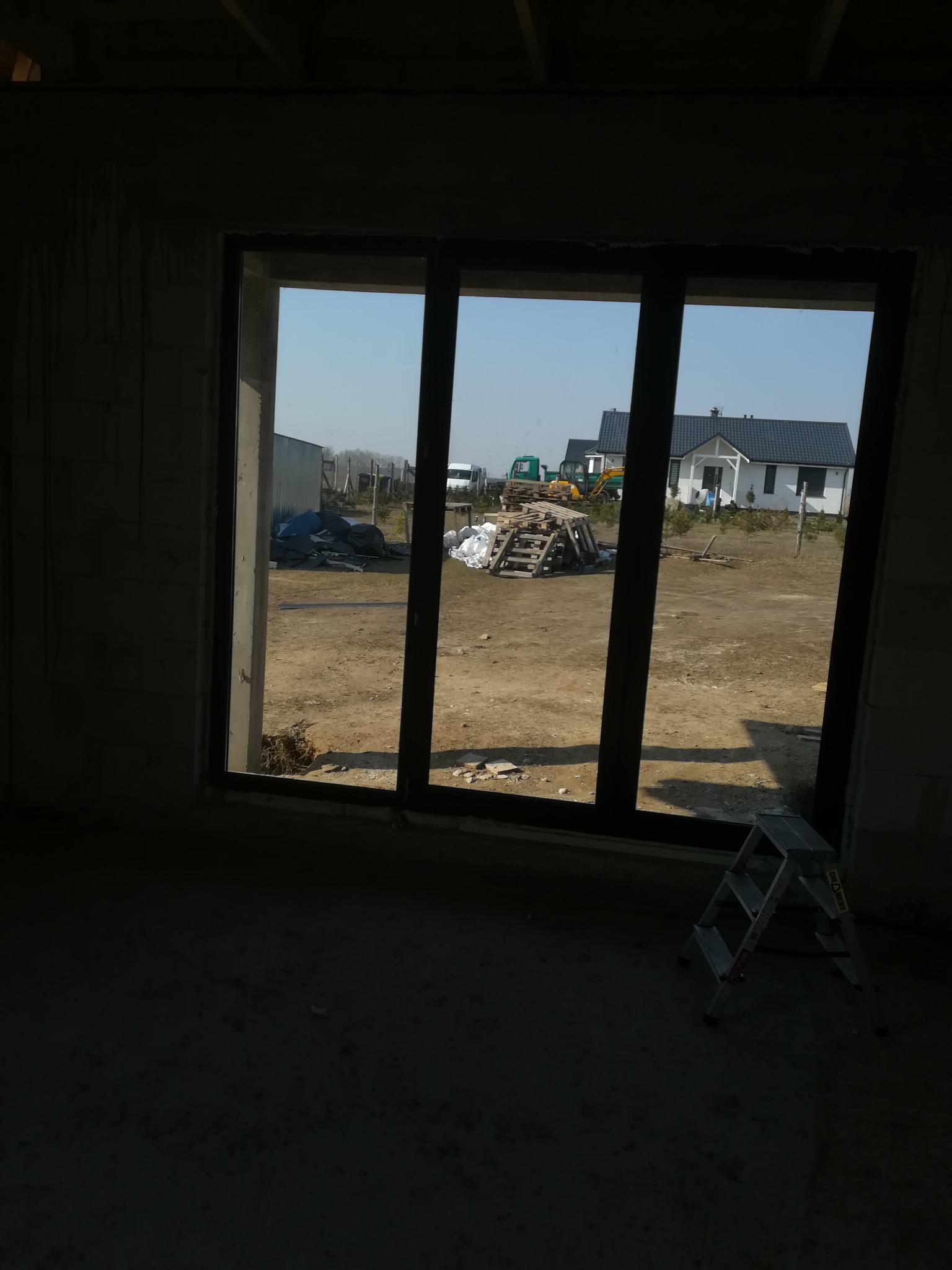 Surowy stan: Czarna rama okienna z widokiem na plac budowy, drabina oparta o ścianę w ciemnym wnętrzu, domy w tle.