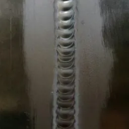 Spoina pachwinowa na metalowej powierzchni, wykonana metodą TIG, widoczne regularne, nachodzące na siebie łuski spawalnicze.