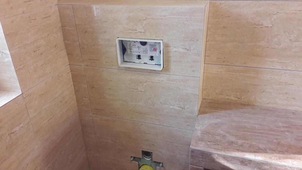 Ściana w łazience wyłożona beżowymi płytkami imitującymi trawertyn, z widoczną wnęką na stelaż podtynkowy WC i fragmentem instalacji kanalizacyjnej w podłodze.